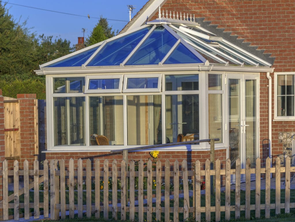A modern glass conservatory with a blue-tinted roof, perfect for conservatory roof repairs, is attached to a brick house and surrounded by a wooden picket fence. Inside the sunlit space with yellow flowers, wicker chairs welcome relaxation.