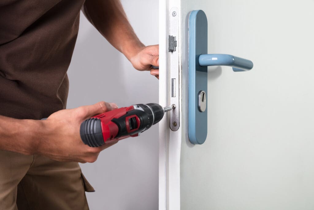 A person using a red and black electric drill to fix the lock mechanism on a white door with a blue handle, demonstrating skills often needed in window repairs and conservatory roof repairs.