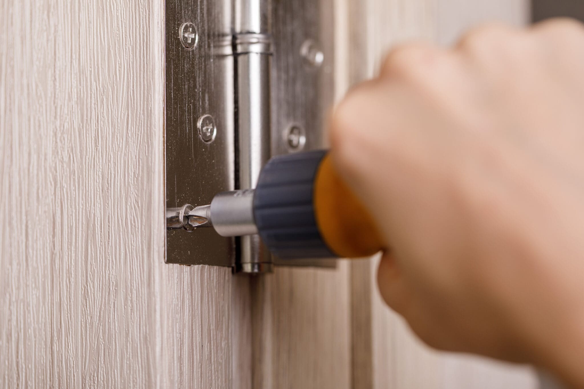 A hand uses a screwdriver to tighten a screw on a metal hinge attached to a light-colored wooden door, showcasing basic window repairs and maintenance.