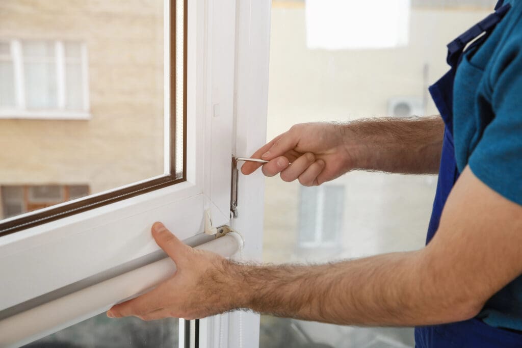 A person in a blue shirt uses a screwdriver to perform window repairs, holding the white window frame with one hand while working indoors near another window.