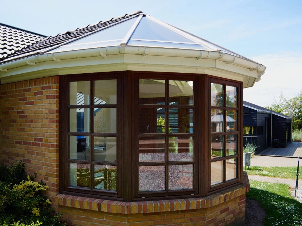 A brick house with a bay window featuring dark brown frames and multiple glass panes, white guttering, and a tiled roof is shown on a sunny day—a great example for those considering expert window repairs or conservatory roof repairs.