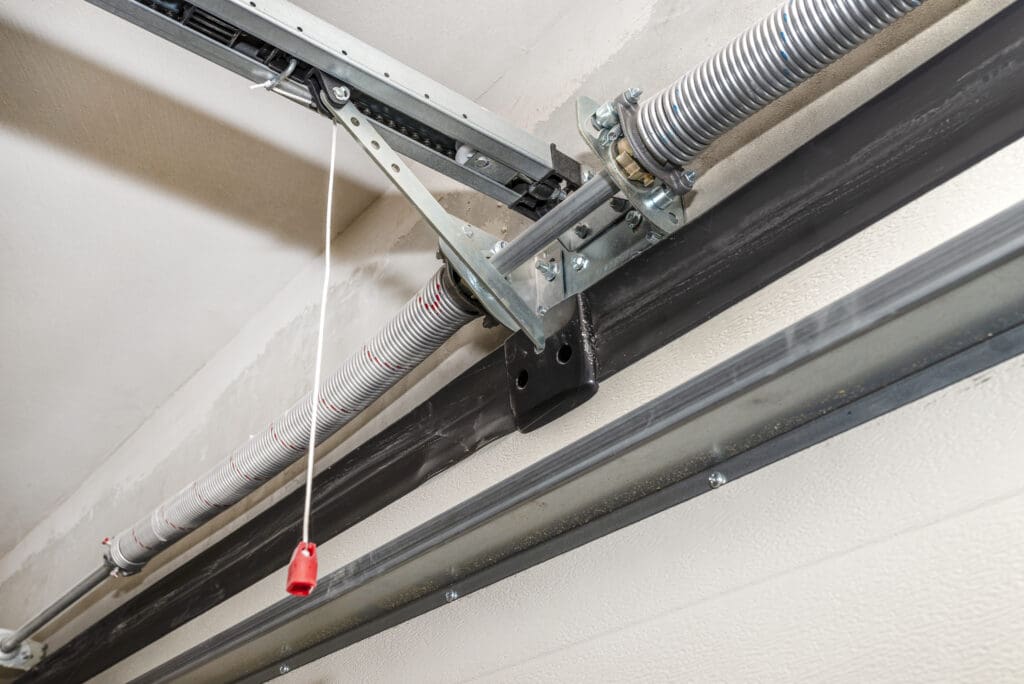 Close-up view of a garage door torsion spring system with a metal rod, brackets, and a red emergency release pull cord—perfect for those needing repairs, whether it's for broken locks or window repairs.