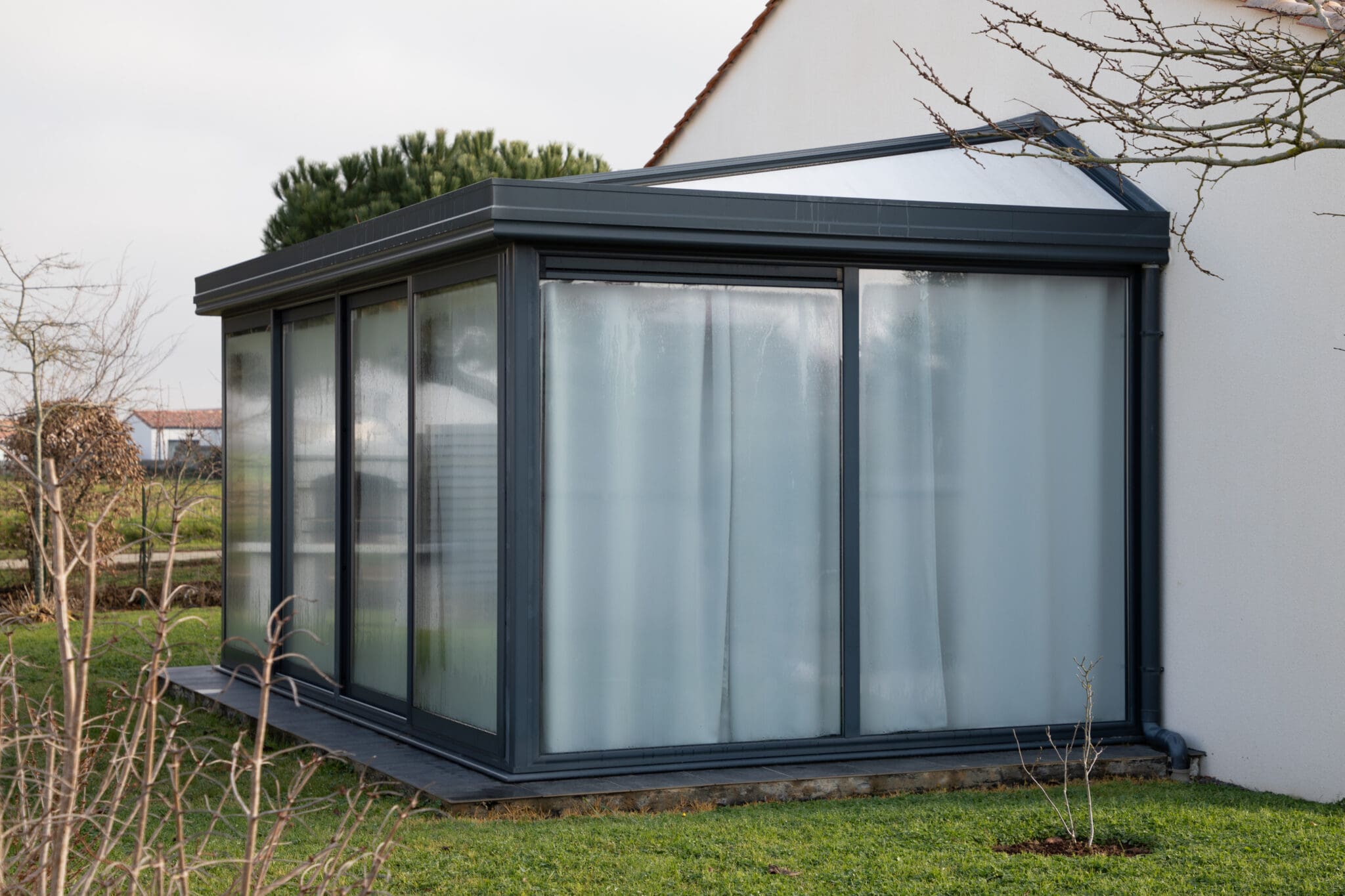 A modern glass sunroom with frosted windows extends from a white house, its conservatory roof repairs expertly handled by a skilled glazier, surrounded by green grass and bare branches on a cloudy day.