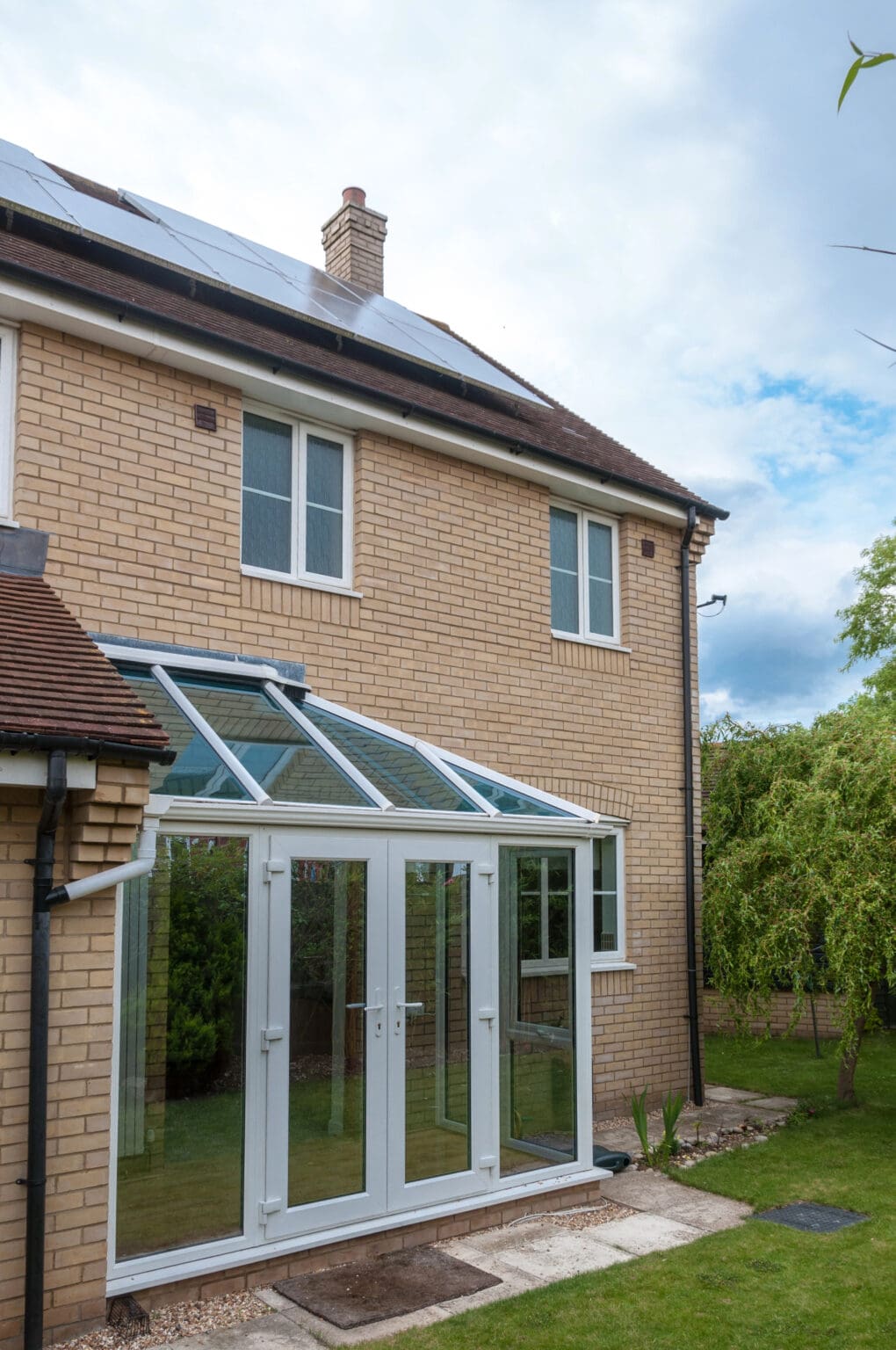 A modern two-story house with light-colored brick walls, a glass conservatory extension featuring double doors, and solar panels on the roof, set next to a garden with grass and trees. Skilled in conservatory roof repairs for lasting comfort.