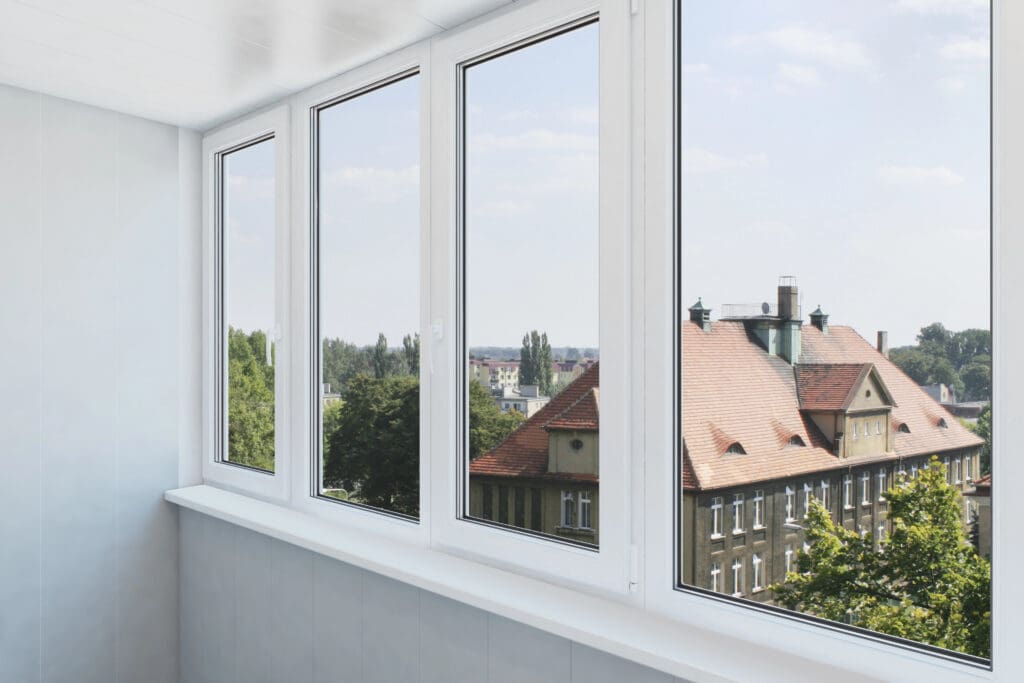 A bright indoor space with large white-framed windows, expertly maintained by a skilled glazier, overlooks rooftops, trees, and a cityscape with clear skies outside.