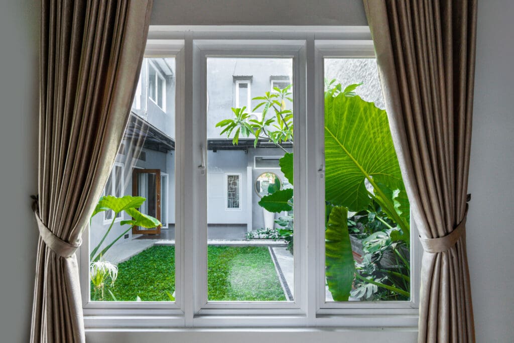 A view through a window with beige curtains, showing a lush green courtyard with large tropical plants, grass, and a modern white building in the background—perfect inspiration for flawless window repairs.