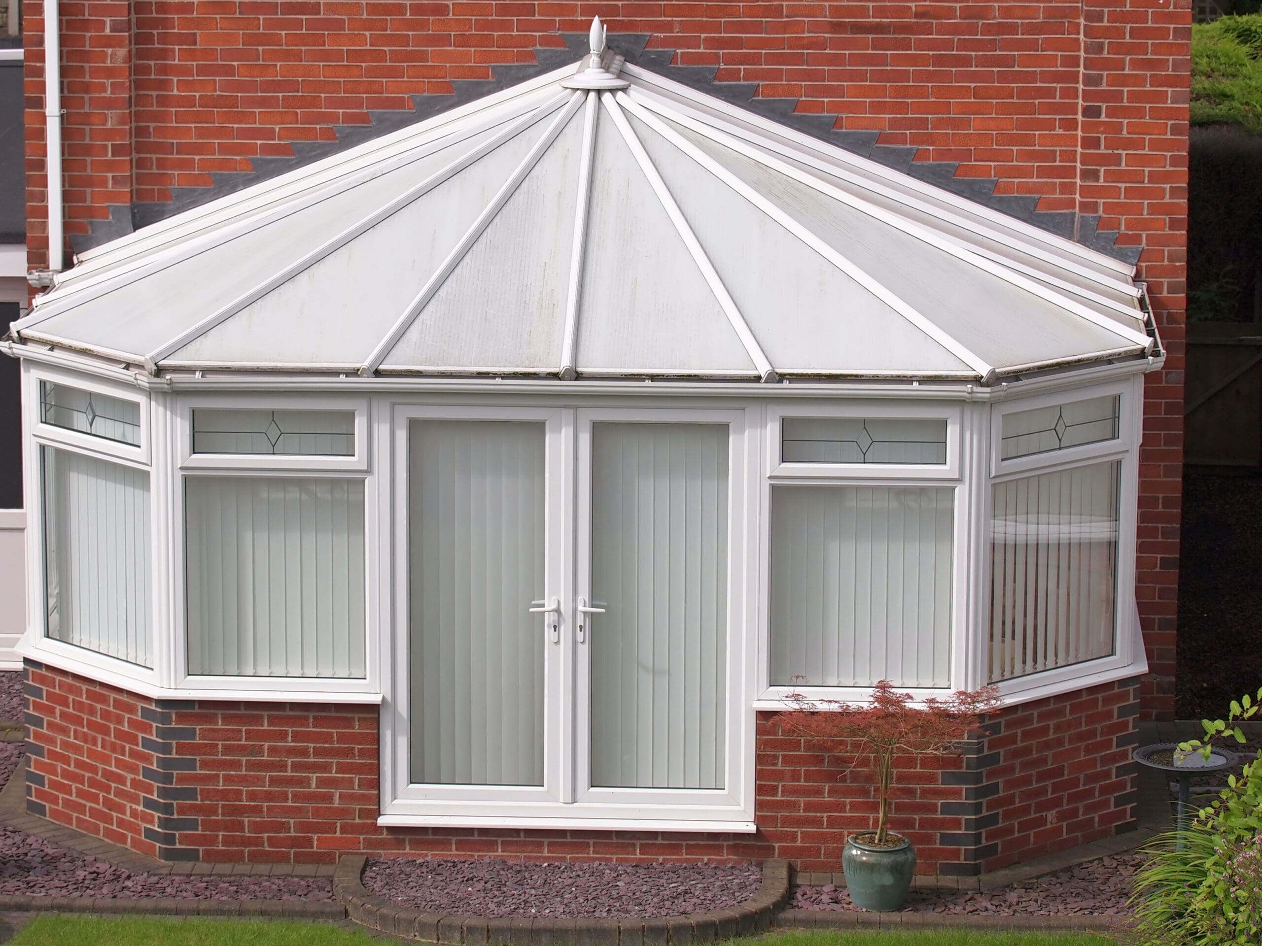 A white-framed glass conservatory with a pointed roof is attached to a red brick house. Vertical blinds cover the windows, and a small potted plant sits on the patio in front—perfect for glazier services or conservatory roof repairs.