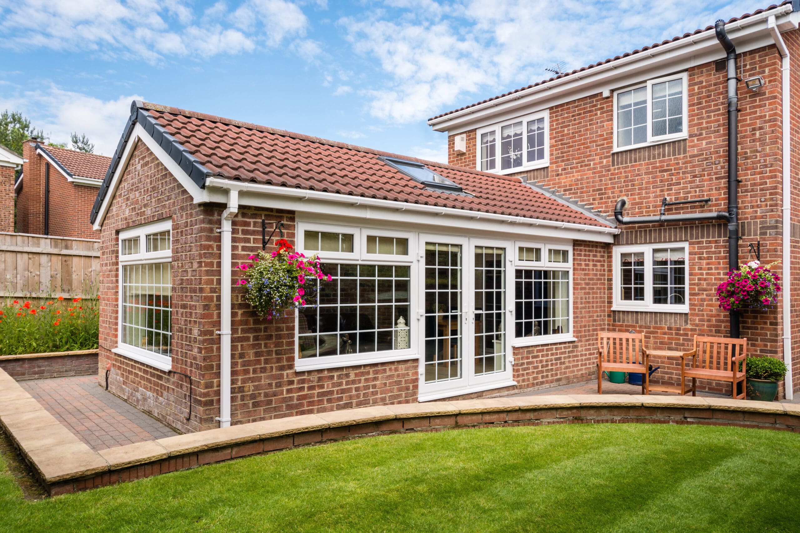 A brick house with a red-tiled roof, large white-framed windows and doors—expertly maintained by skilled glaziers for any window repairs—hanging flower baskets, a wooden bench, and a well-kept green lawn under a partly cloudy sky.